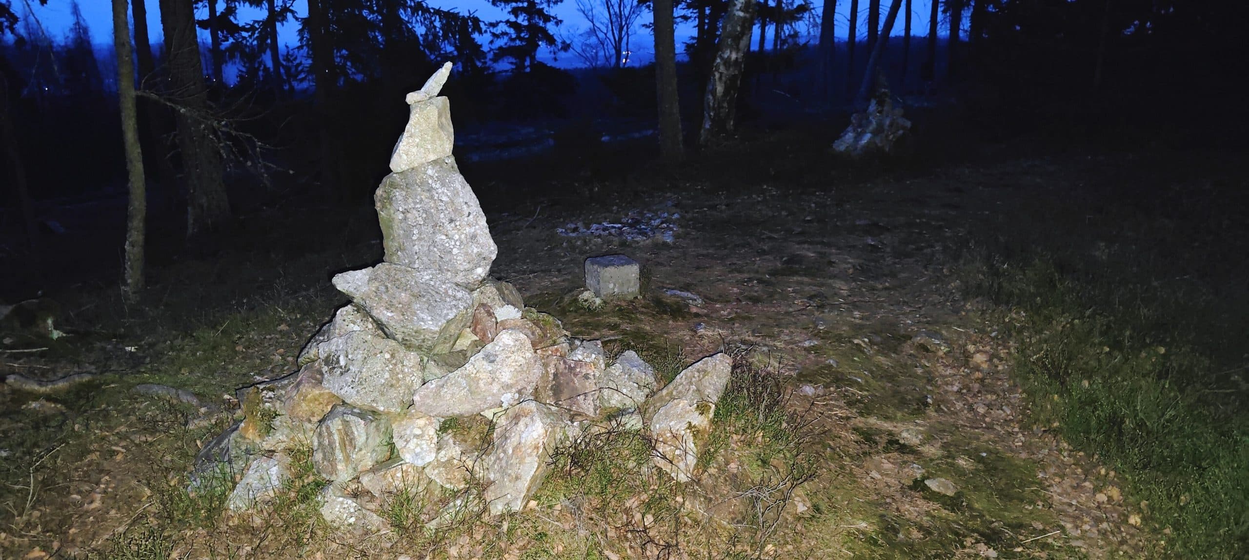 Tall rough stone cairn on a wooded ridge at dusk, with a small concrete block visible beyond it.