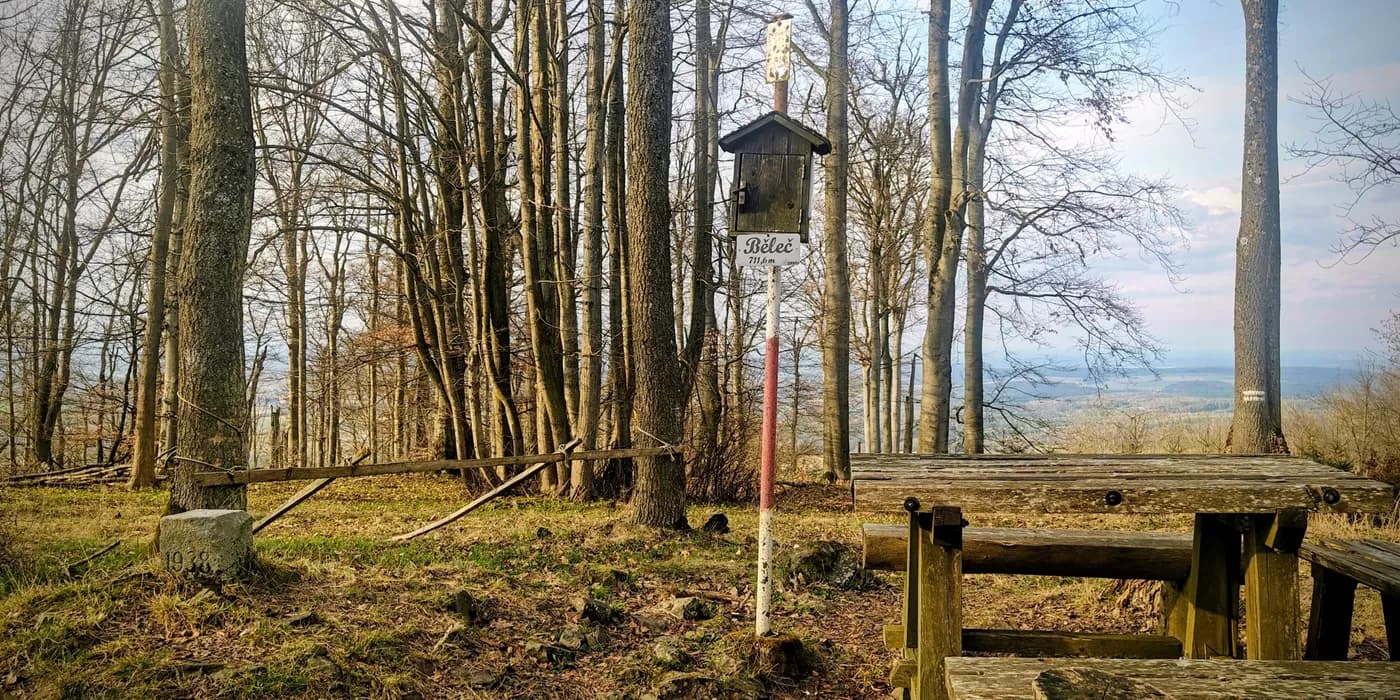 Wooded Běleč summit with a white summit sign, picnic table and concrete marker among leafless trees.