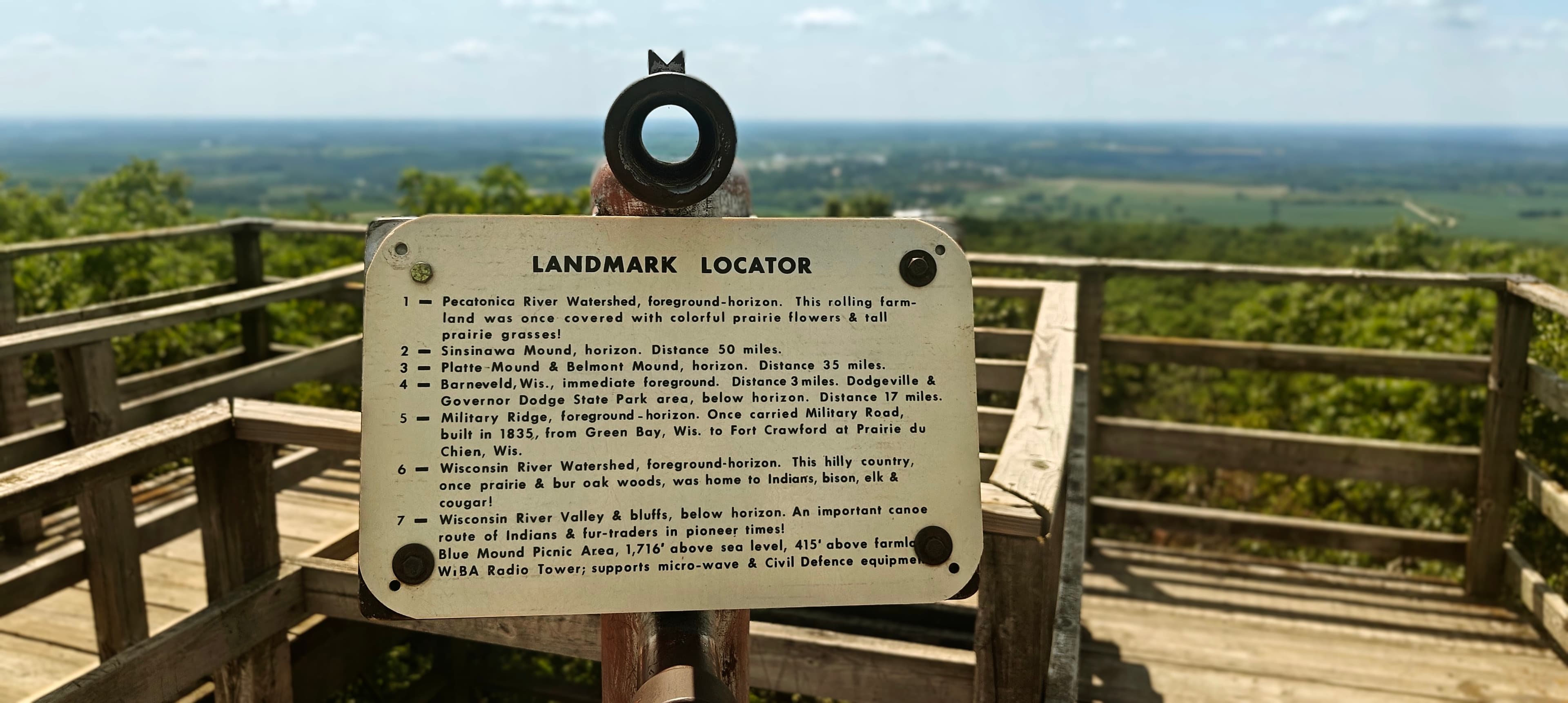 Metal landmark locator on an observation tower deck with wooded countryside beyond.