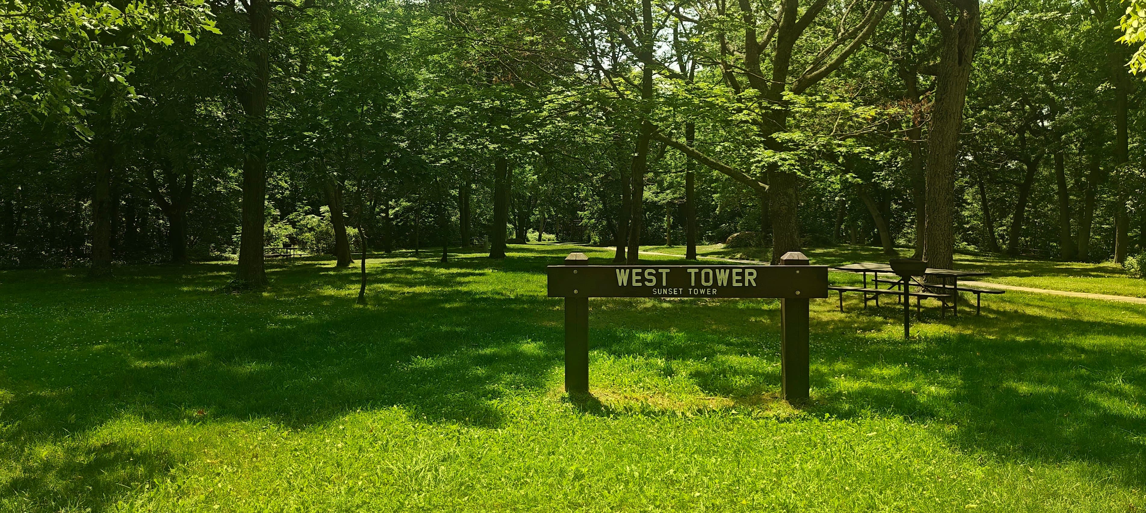 Wooden West Tower sign in a shaded picnic area with tables and trees.