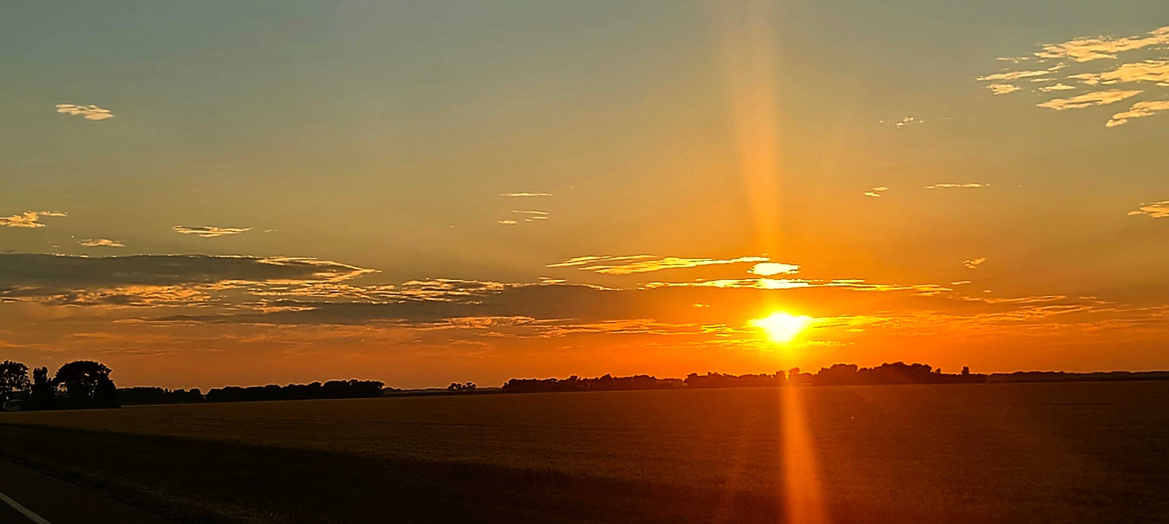Orange sun setting over flat fields beside a road, with scattered clouds glowing in the evening sky.