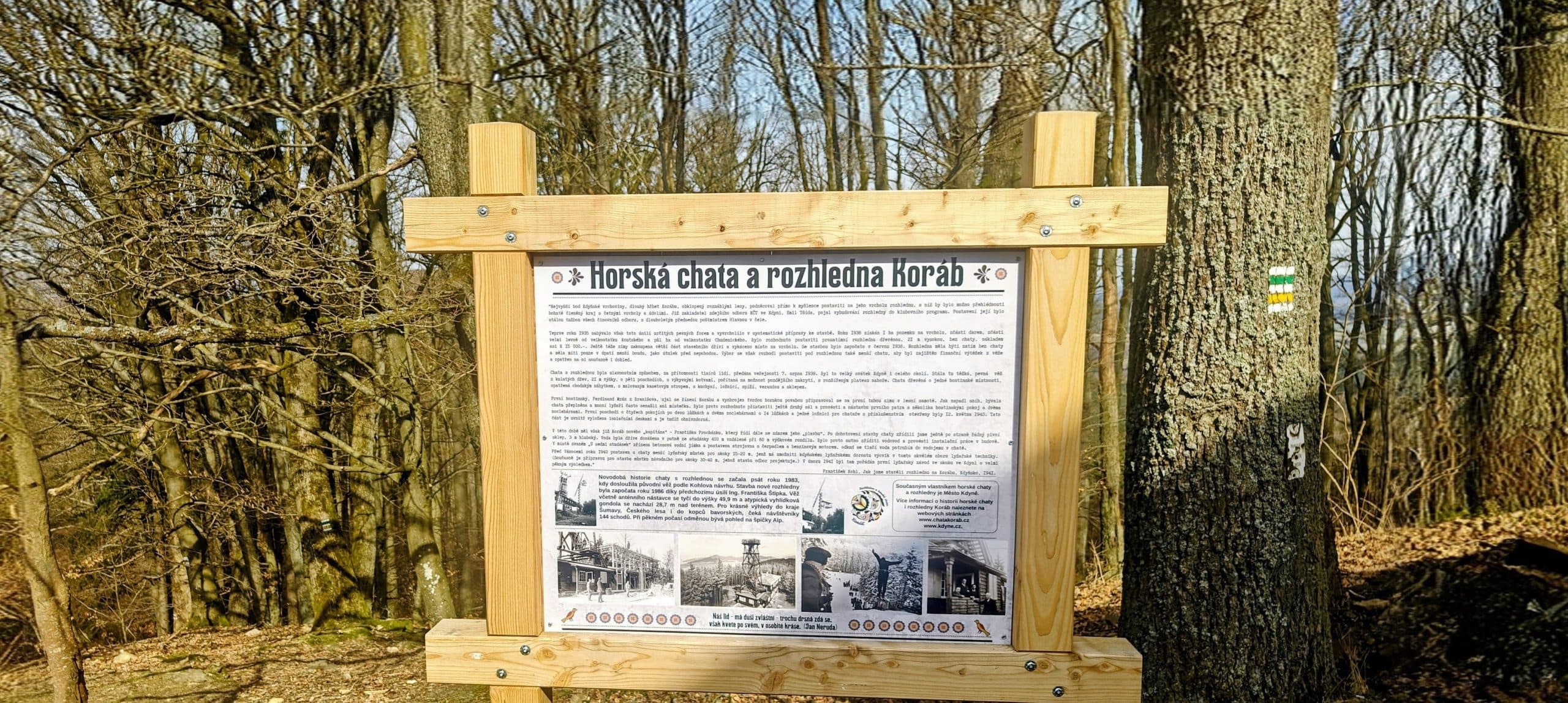 Wooden Koráb hut and lookout history board beside a marked tree in the summit forest.