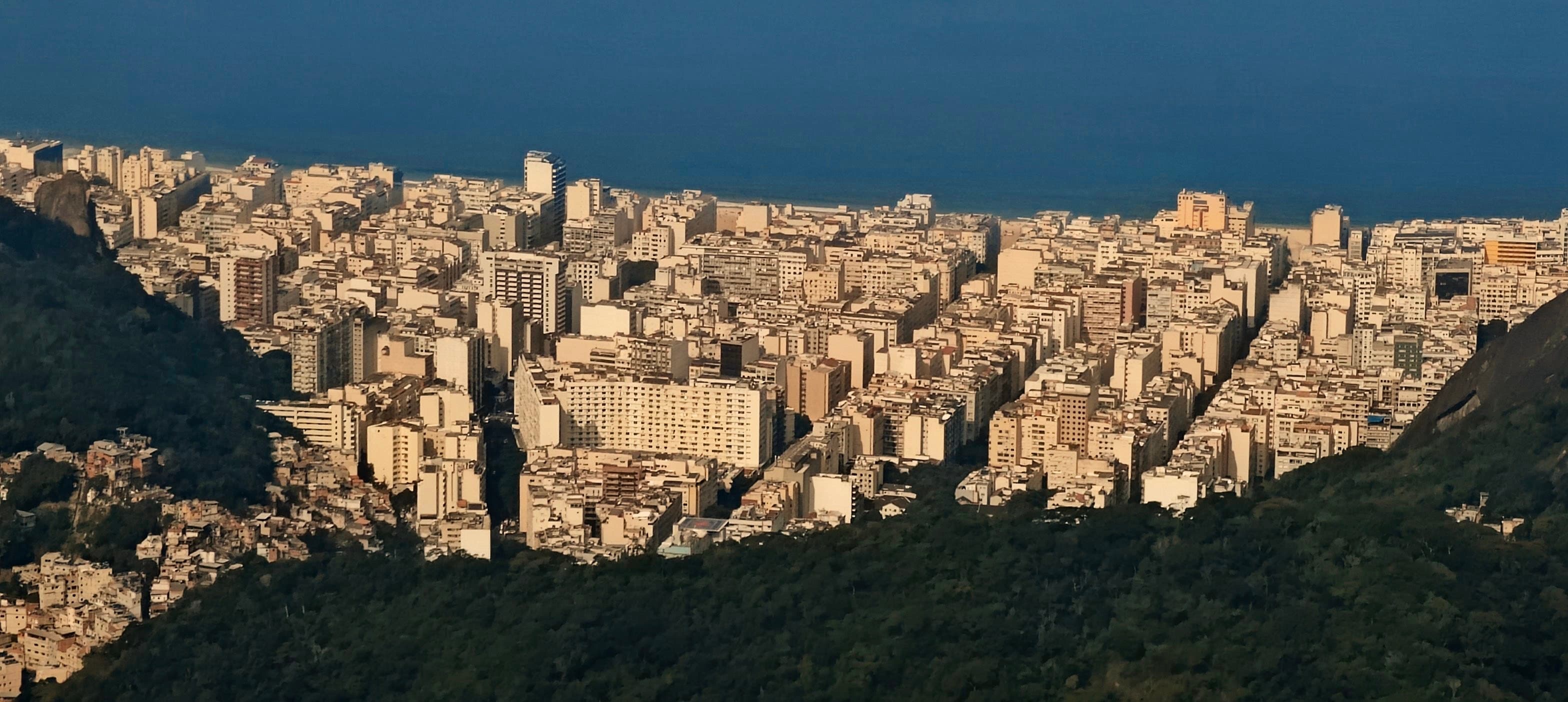 Dense Rio neighbourhoods framed by dark forested slopes with the Atlantic Ocean beyond.
