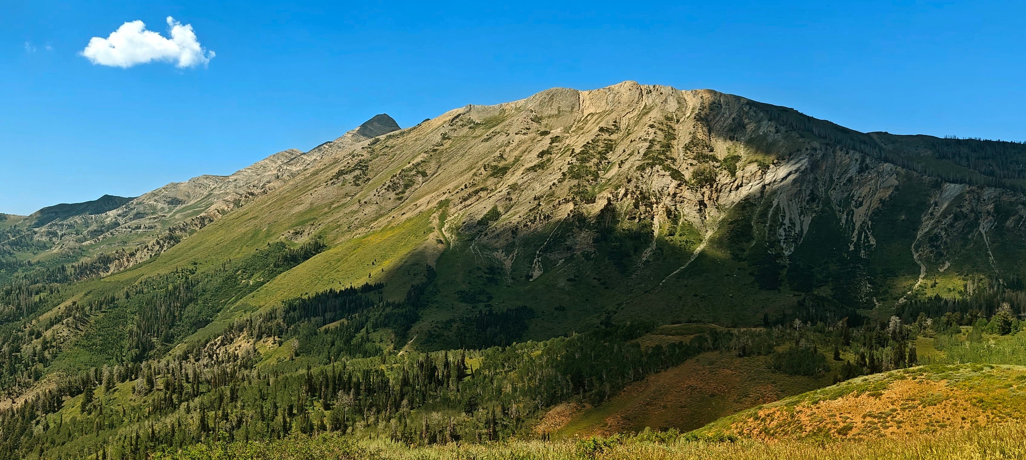 A pale rocky mountain ridge rising above green slopes under a blue sky.