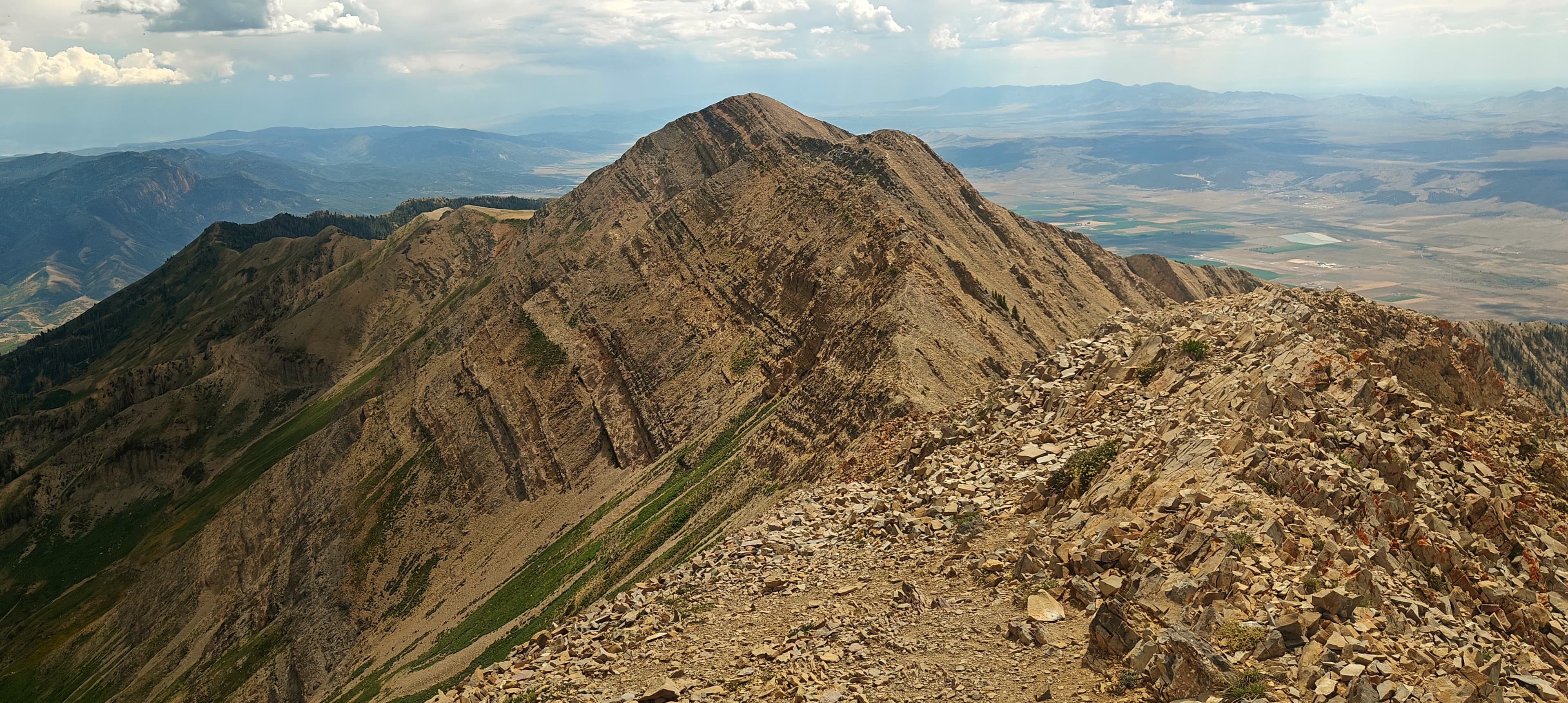 A rugged rocky ridge with steep folded slopes and distant valleys under a partly cloudy sky.