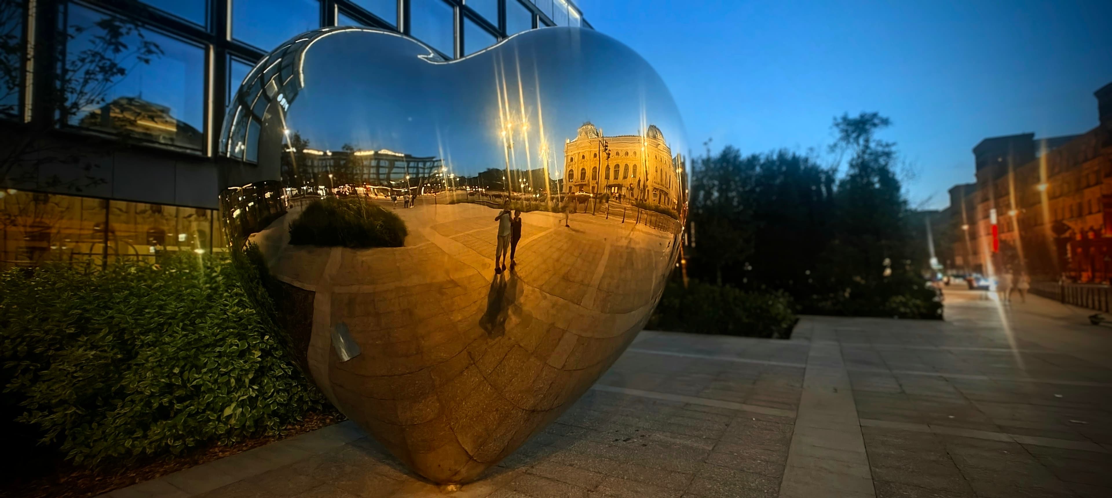 Large polished heart sculpture reflecting buildings and lights beside Manufaktura in Łódź.