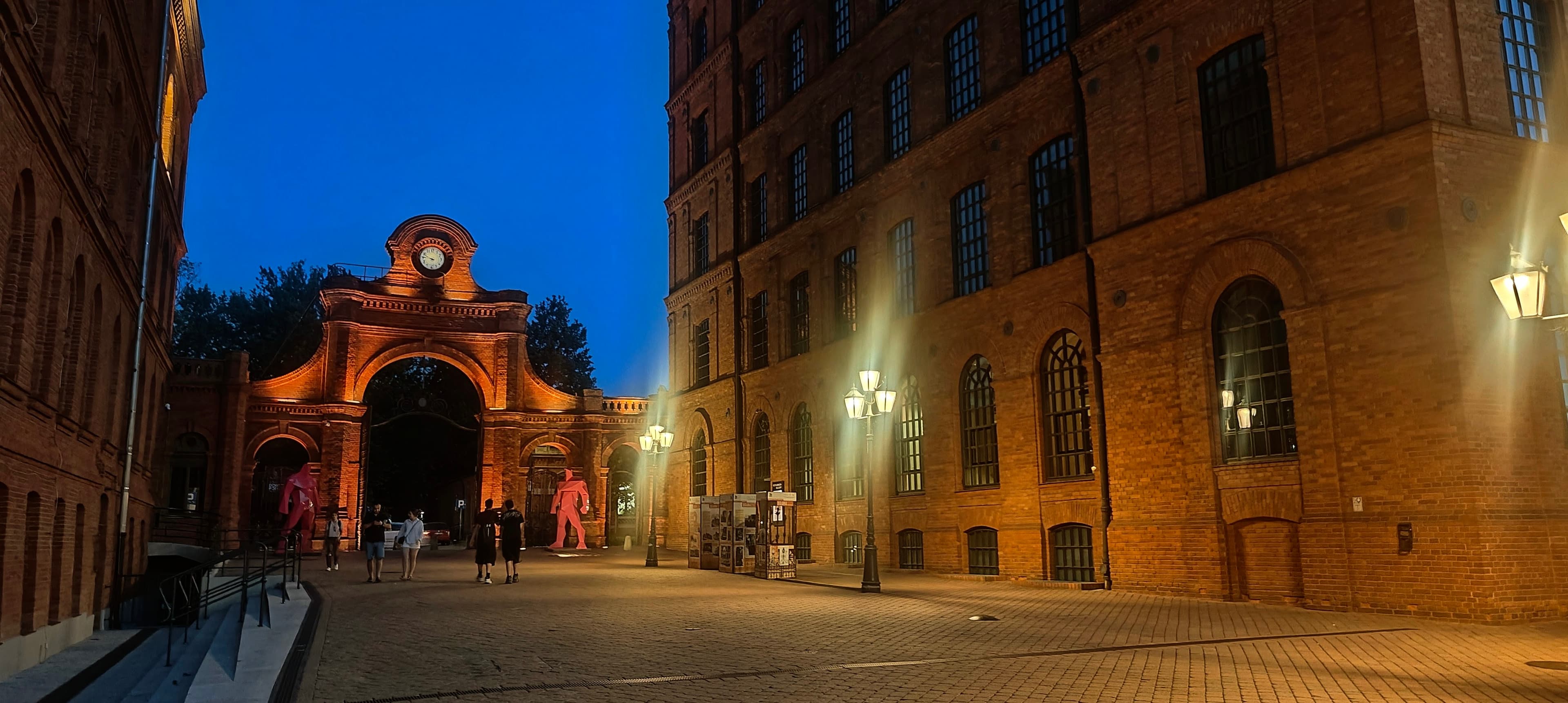 Red-brick arched Manufaktura gate illuminated at blue hour between former factory buildings.