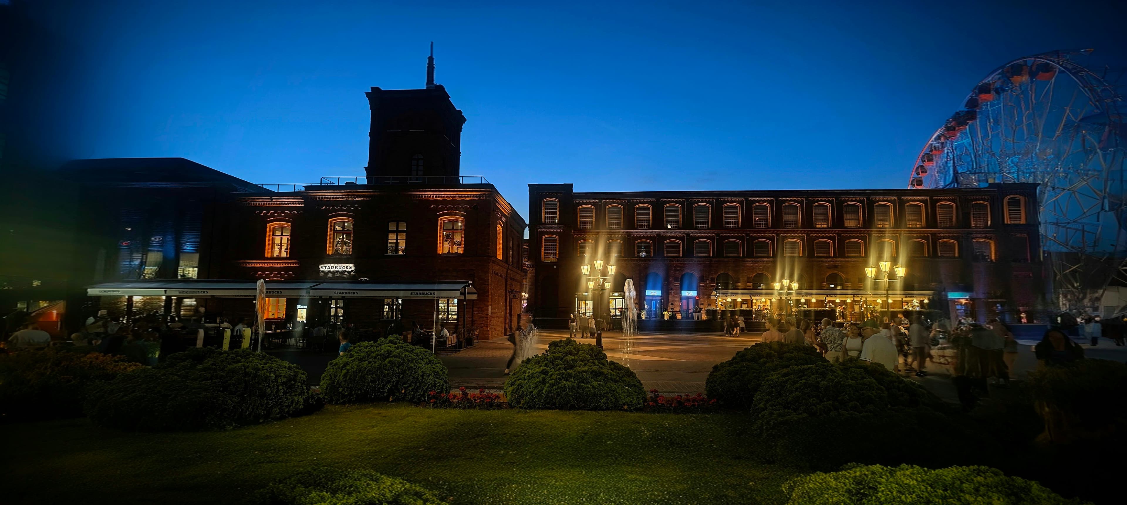 Manufaktura courtyard at blue hour with red-brick factory buildings, gardens and a Ferris wheel.