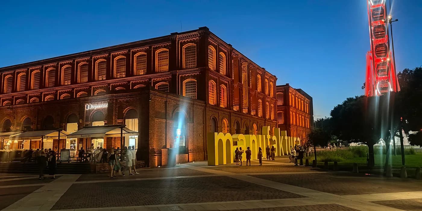 Illuminated red-brick Manufaktura building at blue hour with yellow sign and Ferris wheel.