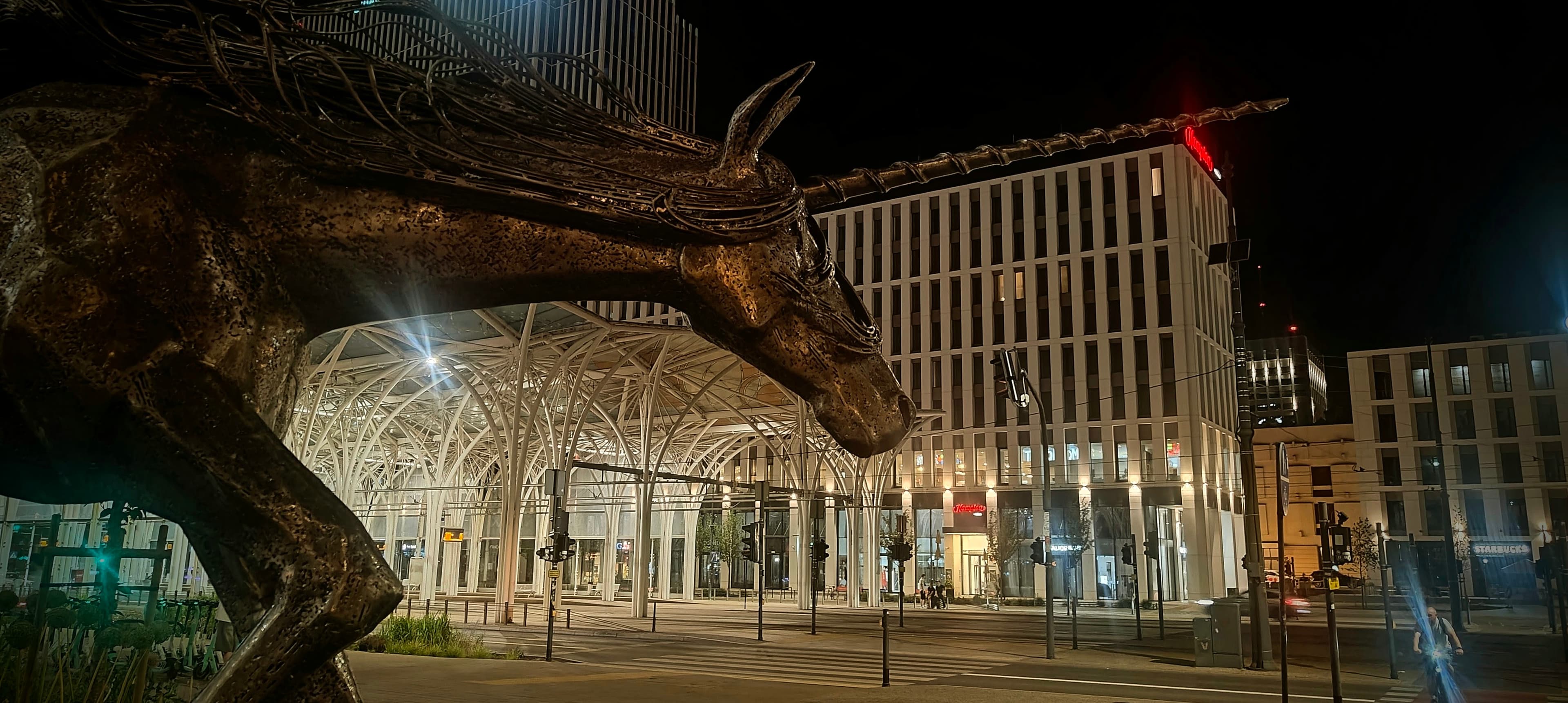 Metal Unicorn Monument in front of the illuminated Piotrkowska Centrum tram shelter at night.