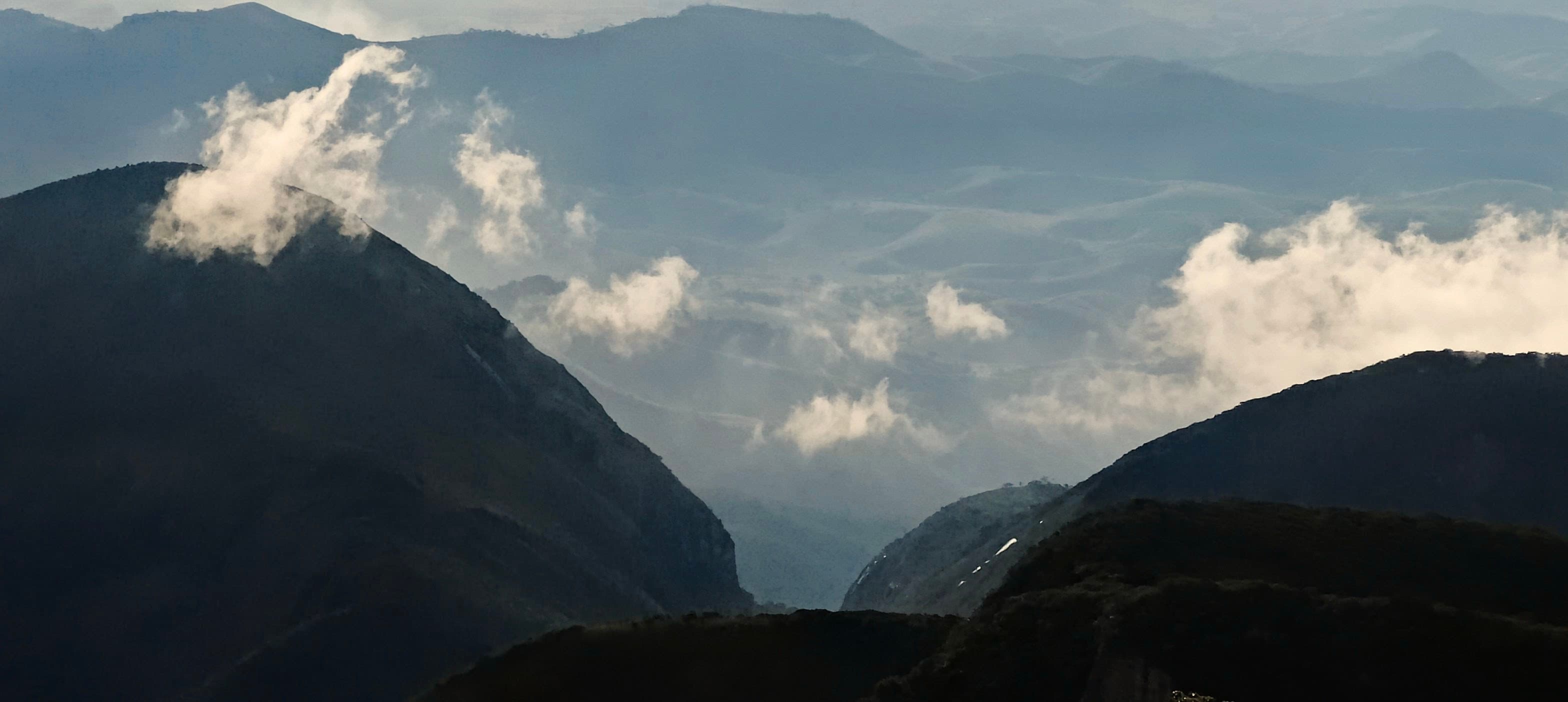 A dark steep mountain peak framed by drifting white clouds and distant ridges.