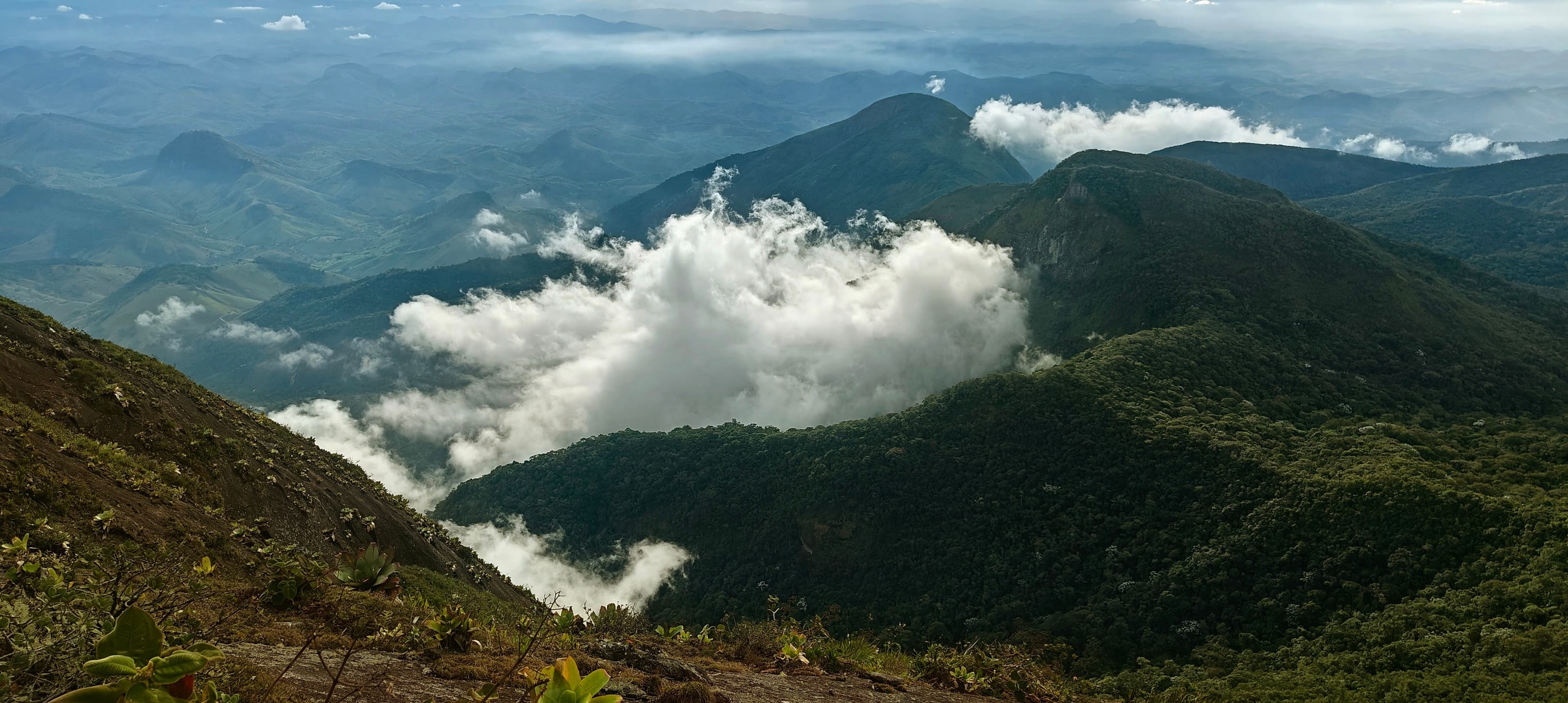 Low white clouds rising through a deep green mountain valley below the summit.