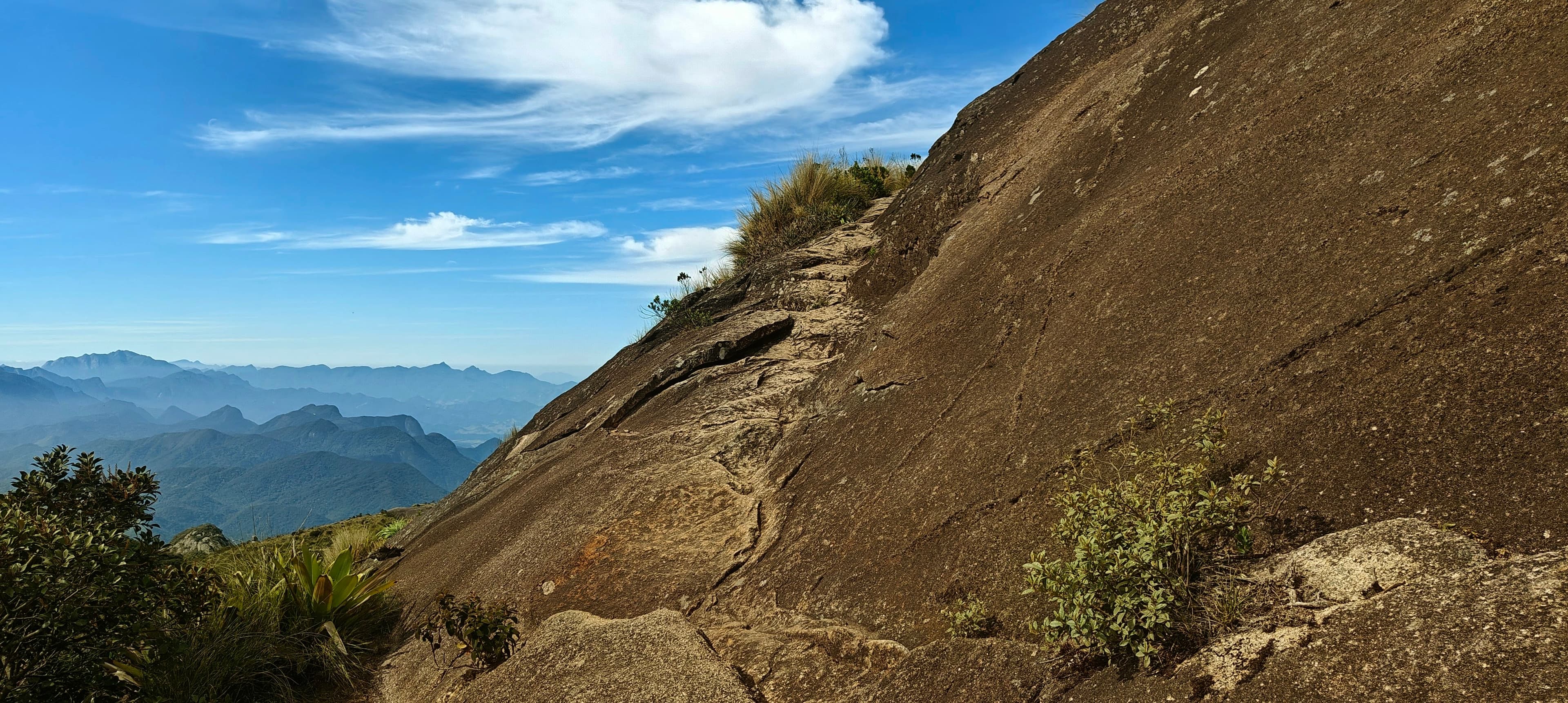A narrow path crossing beside a steep granite slab with distant mountains beyond.