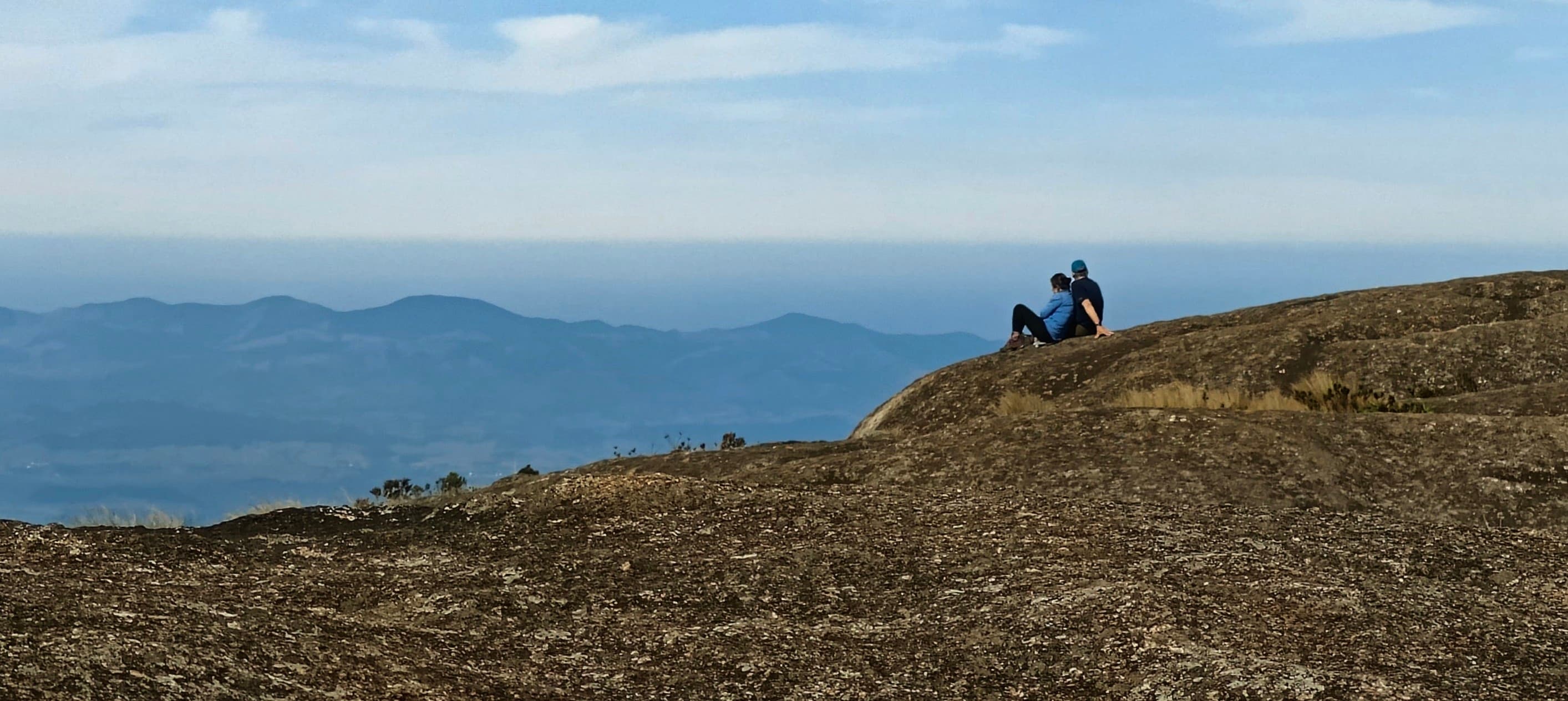 Lihua and Michael sitting together on a broad rock slab near the summit of Pedra do Sino, looking towards Rio and Guanabara Bay.