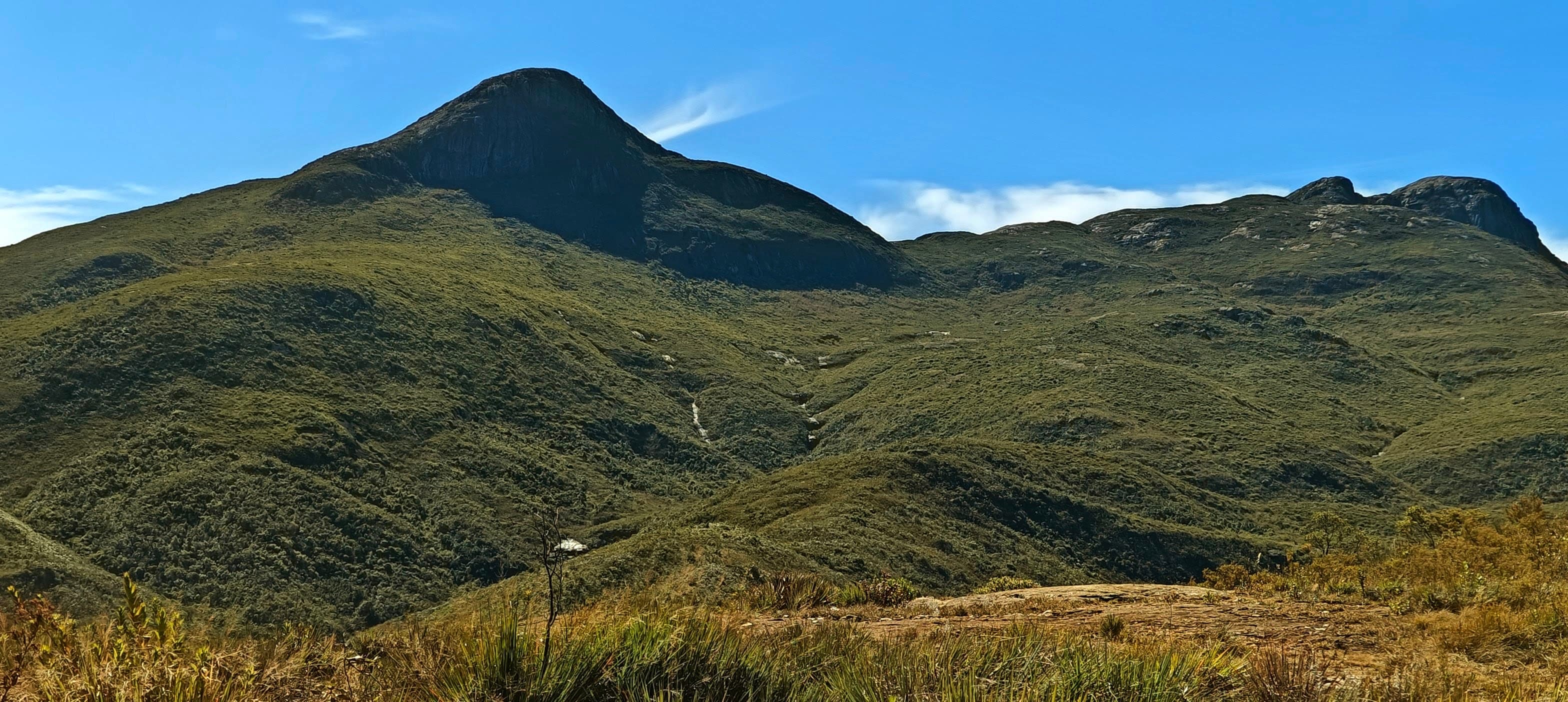 Rounded high peak and open grassland in the Caparaó massif under a clear blue sky.