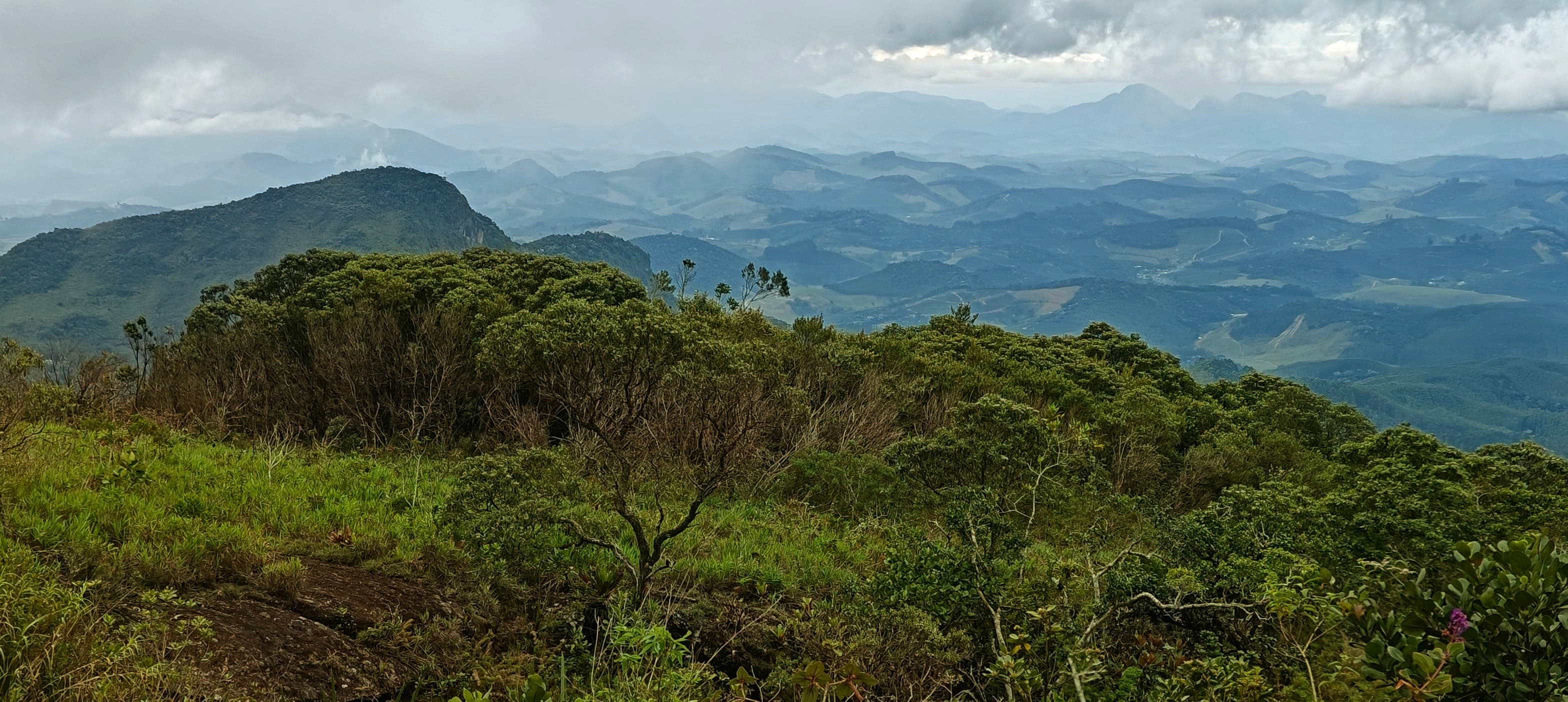 Green ridge vegetation dropping towards layered hills beneath a cloudy sky.