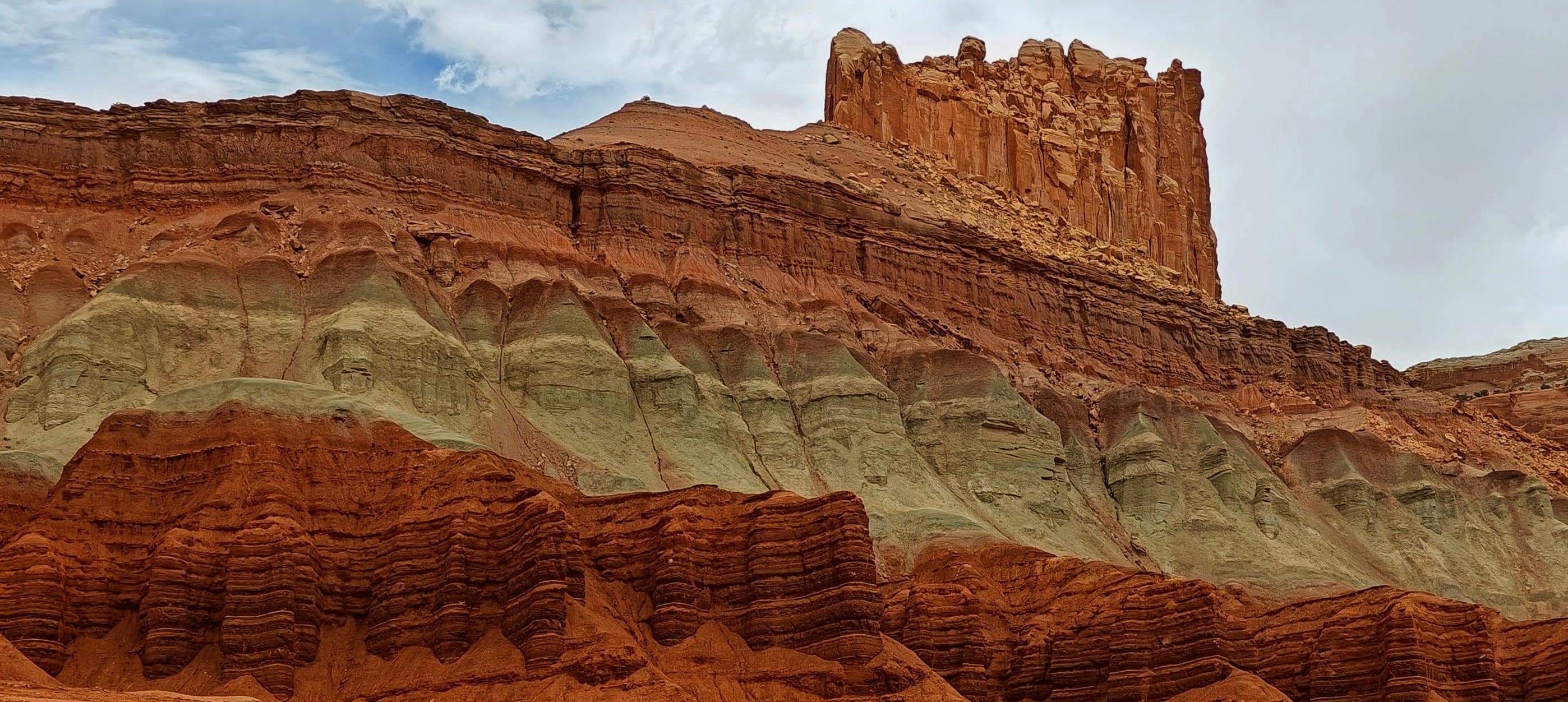 Red and pale green-gray sedimentary rock layers exposed beneath a jagged sandstone ridge in Capitol Reef National Park.