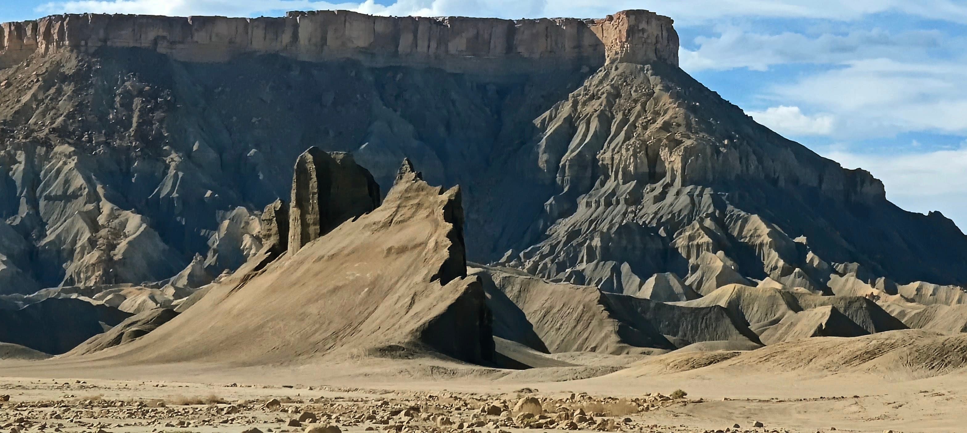 Factory Butte rising behind sharply eroded gray Mancos Shale ridges and a narrow rock pinnacle in Utah.