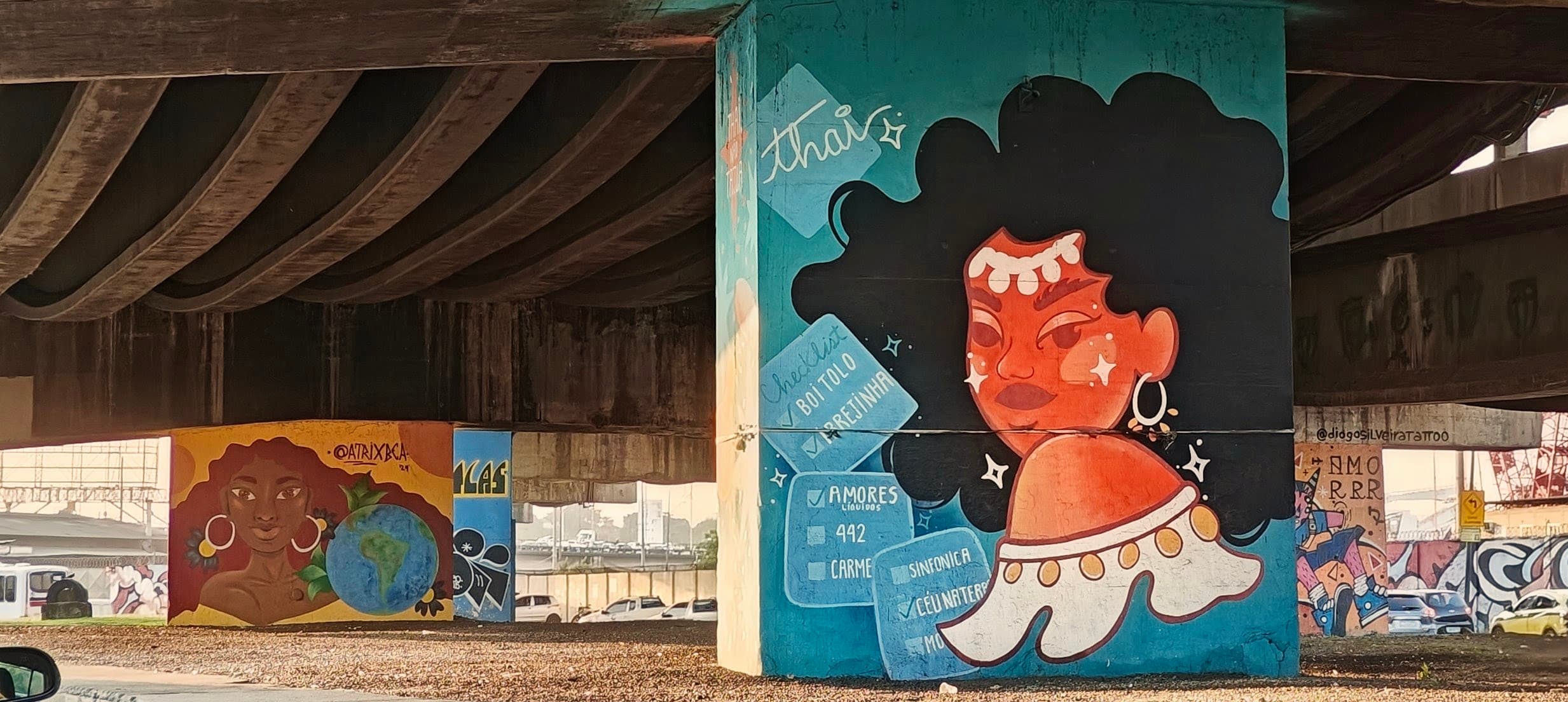 A long wall beneath a concrete viaduct is covered with colourful murals and graffiti.