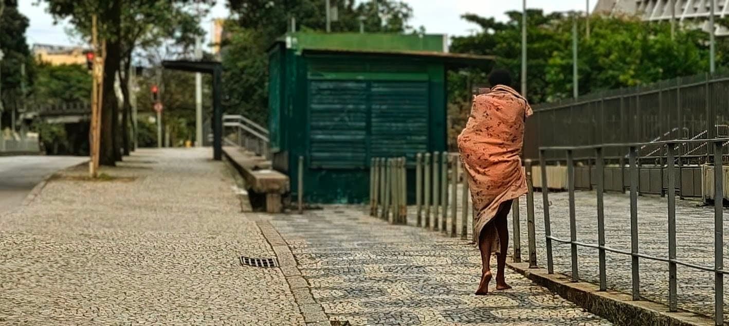 A barefoot homeless person wrapped in orange fabric walks along a broad pavement in central Rio.