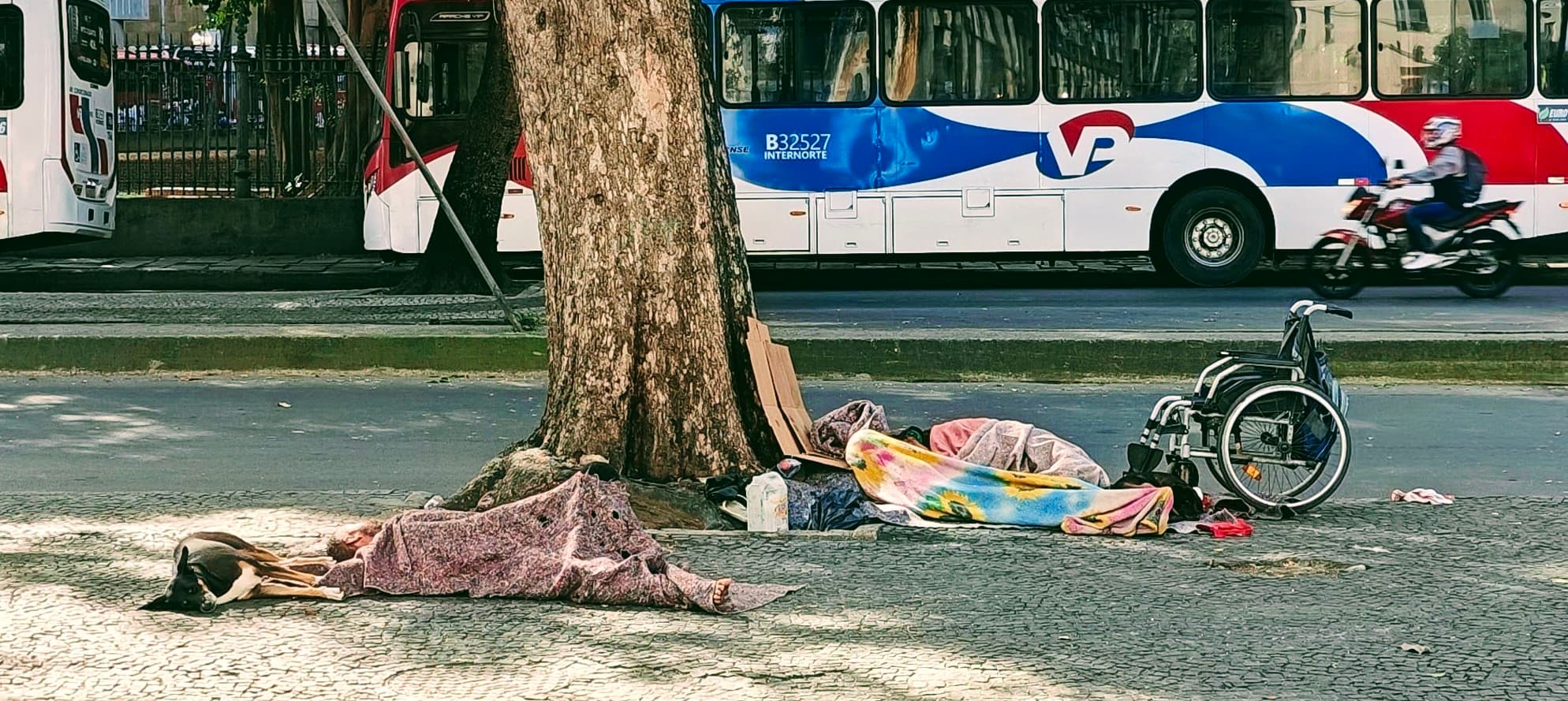 A homeless person rests beside a tree and wheelchair on a pavement in central Rio.