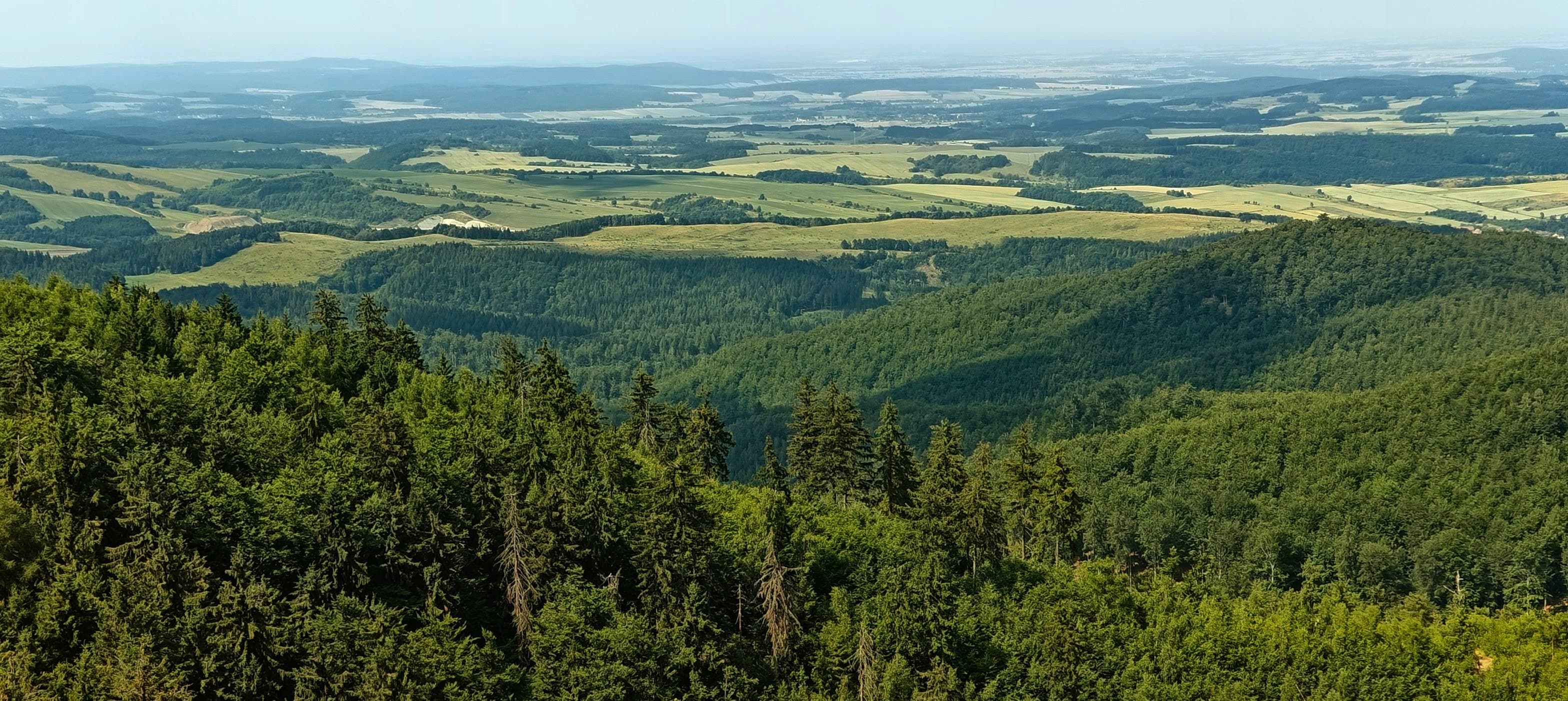 Rolling forested hills and patchwork farmland extending toward a hazy horizon from Trójgarb.