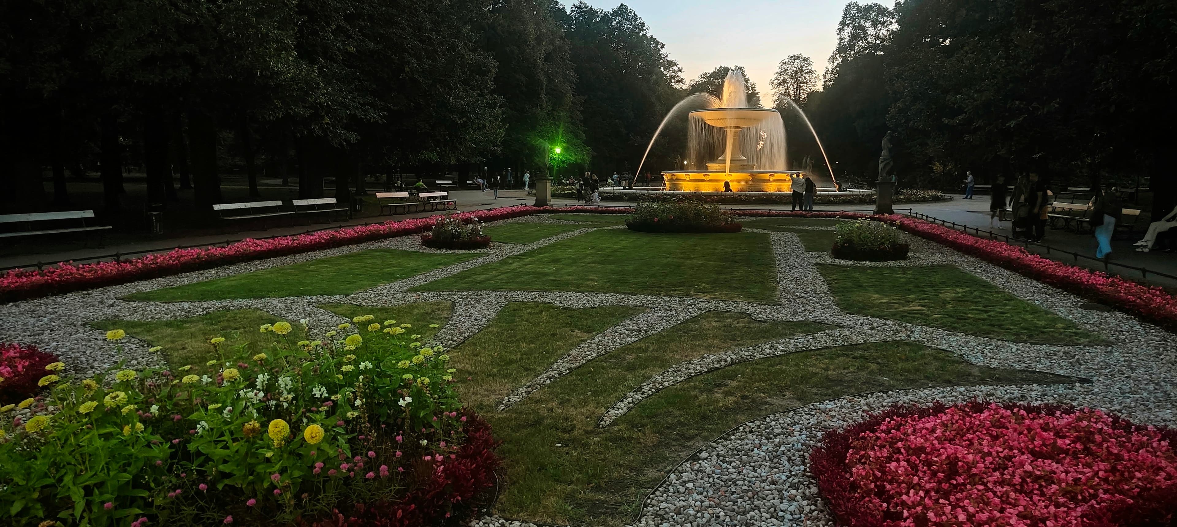 Illuminated fountain beyond geometric flower beds in the Saxon Garden in Warsaw at dusk.