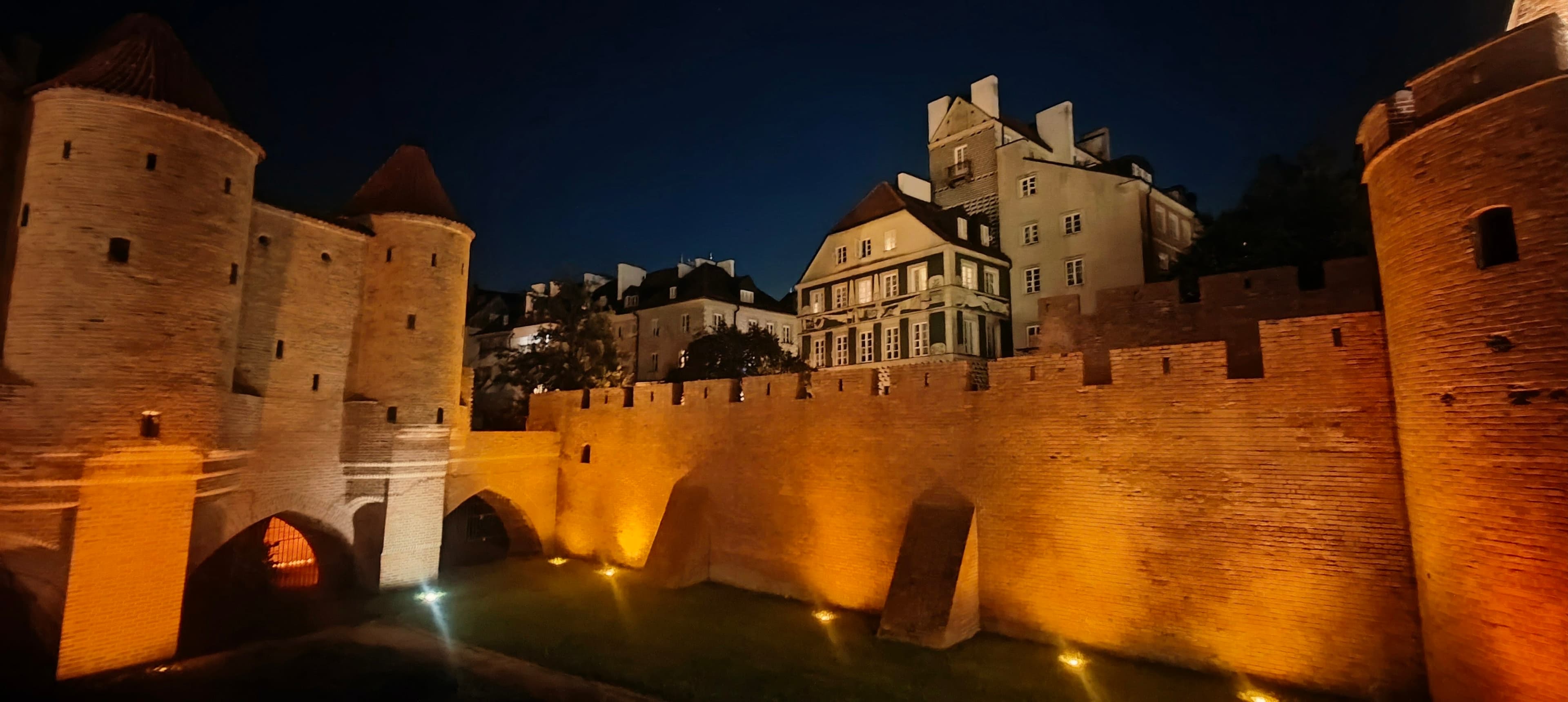 Illuminated brick defensive walls and round towers around Warsaw Old Town at night.