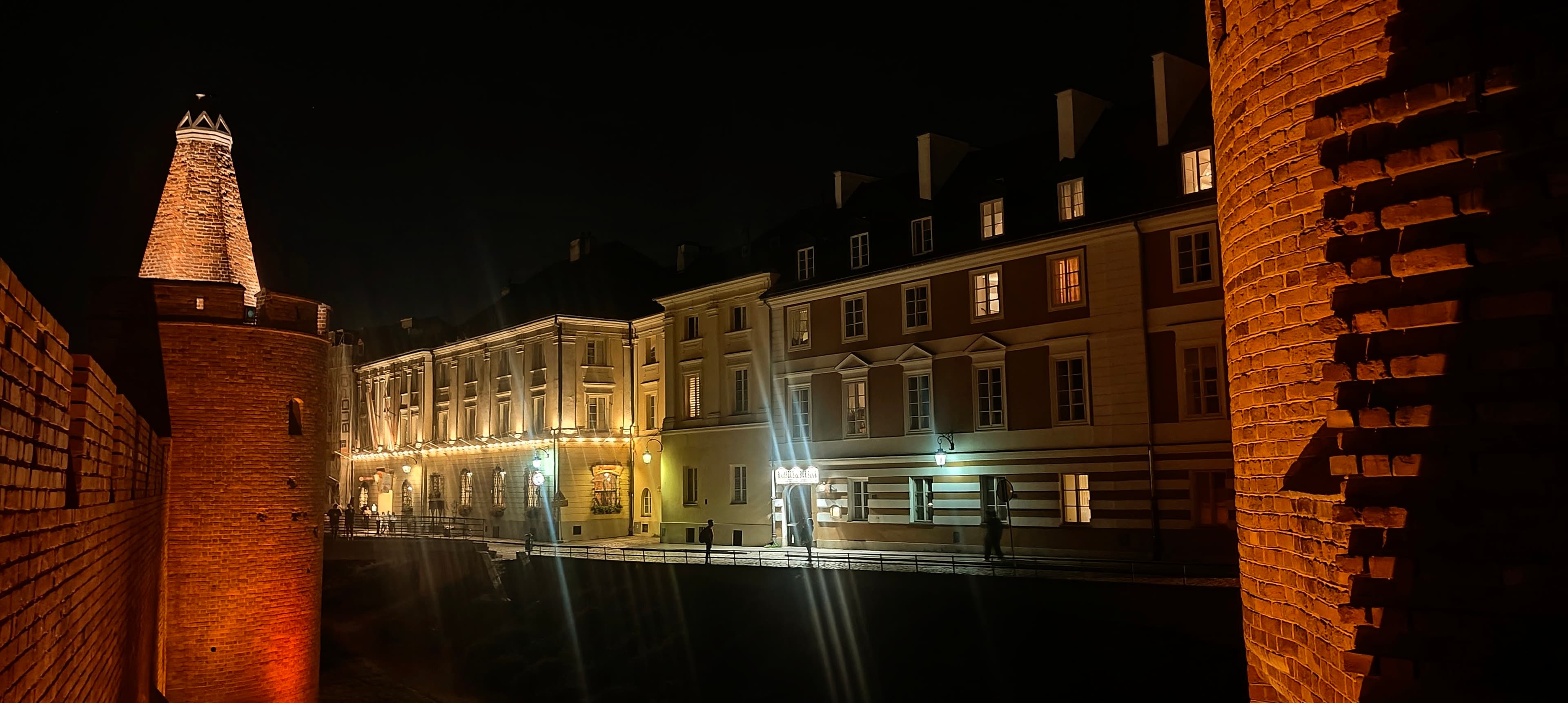 Brick tower and defensive wall framing illuminated townhouses in Warsaw Old Town at night.
