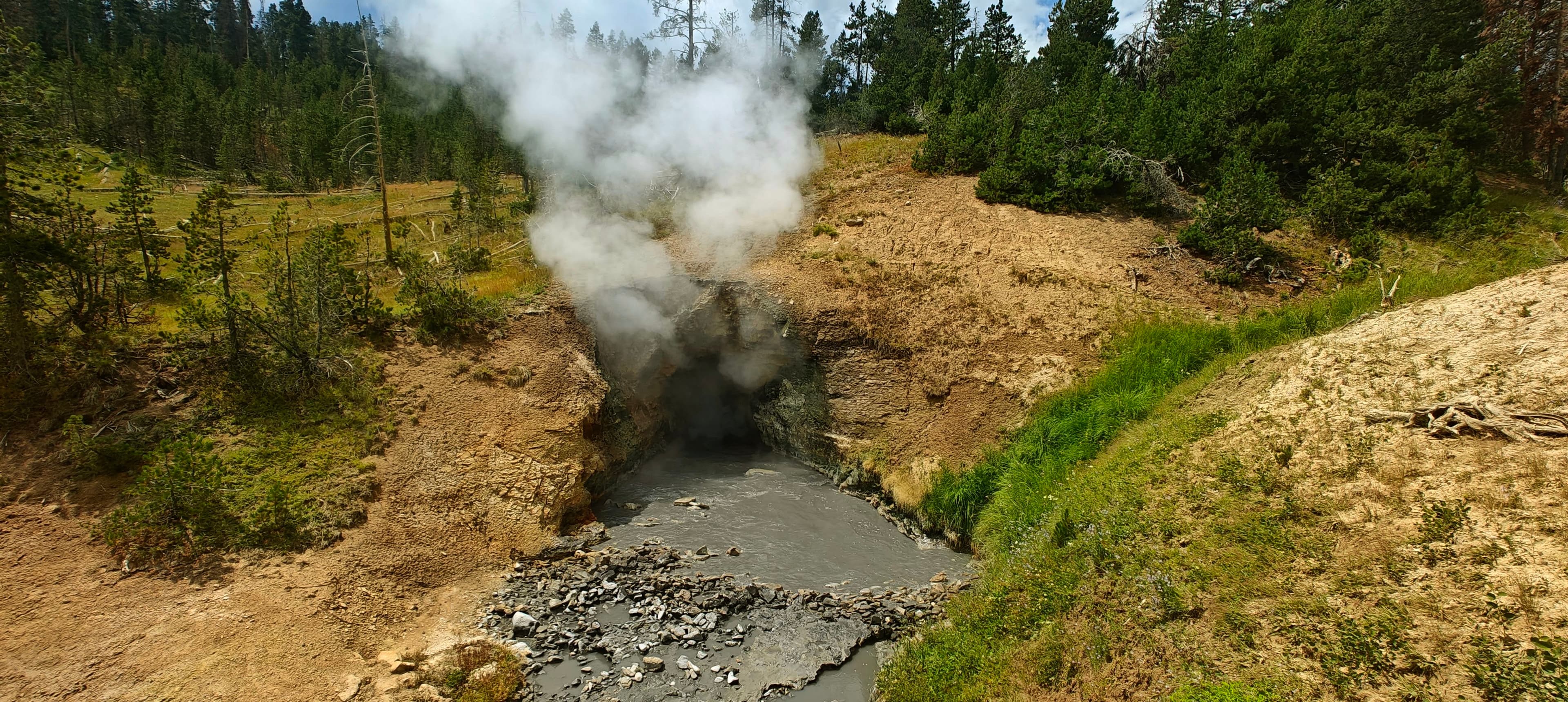 Steam rises from a narrow grey mud channel cut through ochre geothermal ground.