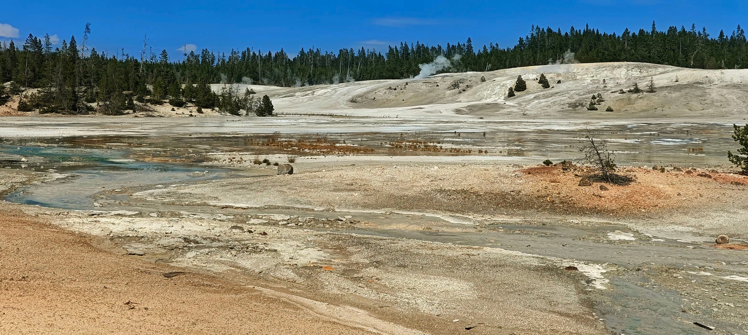 Pale mineral ground and scattered pools stretch across a broad basin under blue sky.