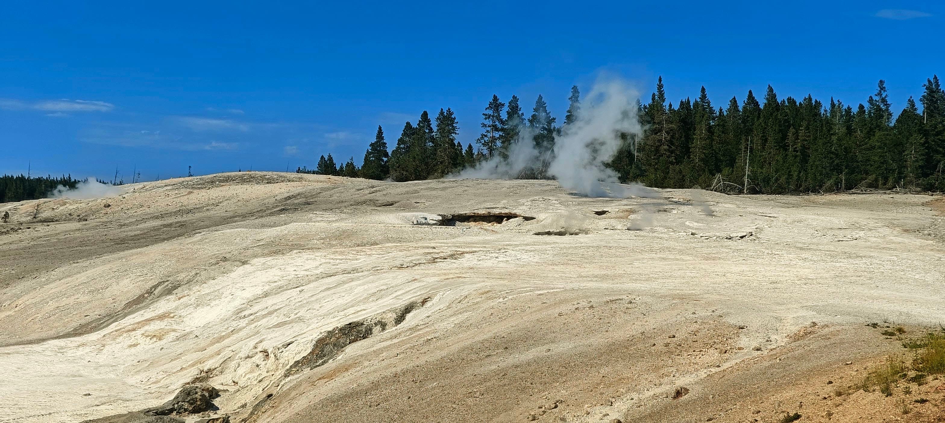 Steam vents punctuate a smooth pale geothermal ridge beneath a deep blue sky.