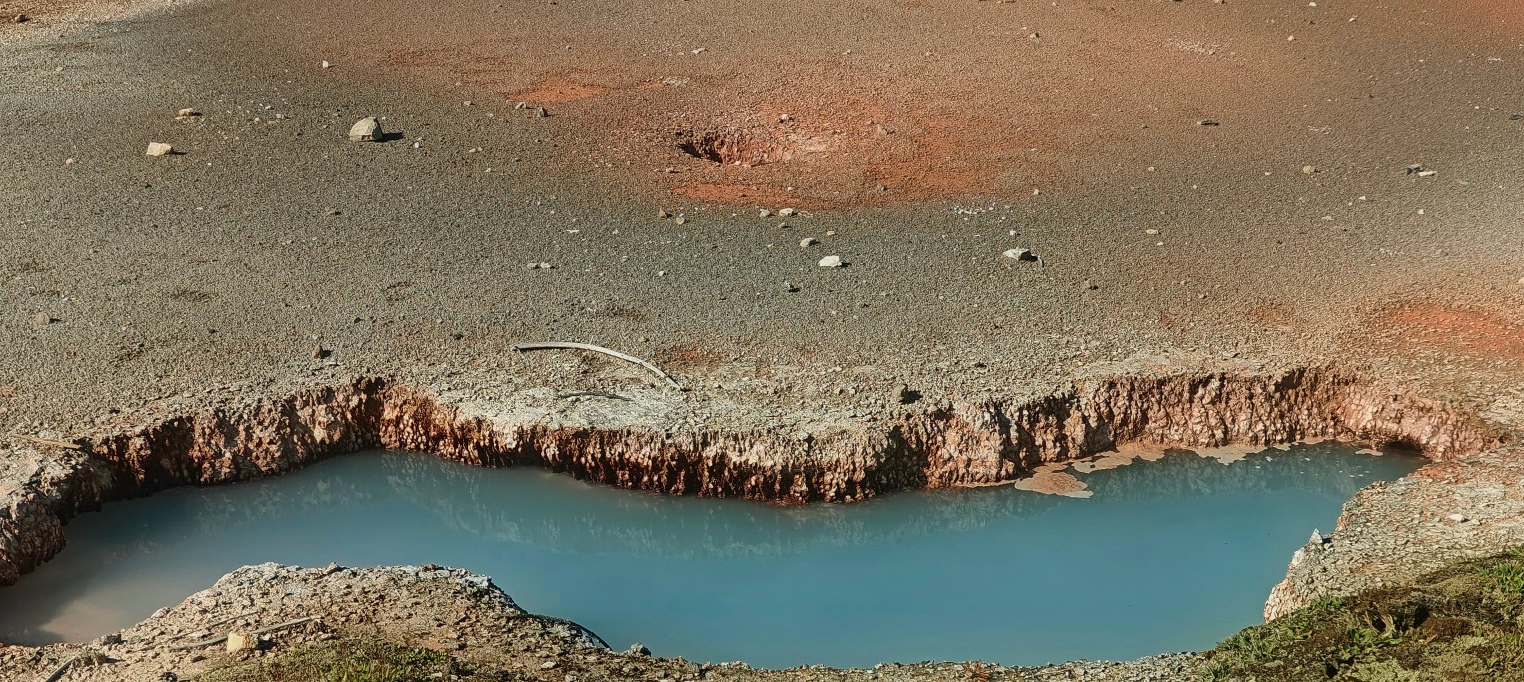 A vivid red-orange microbial mat borders blue thermal water beneath drifting steam.
