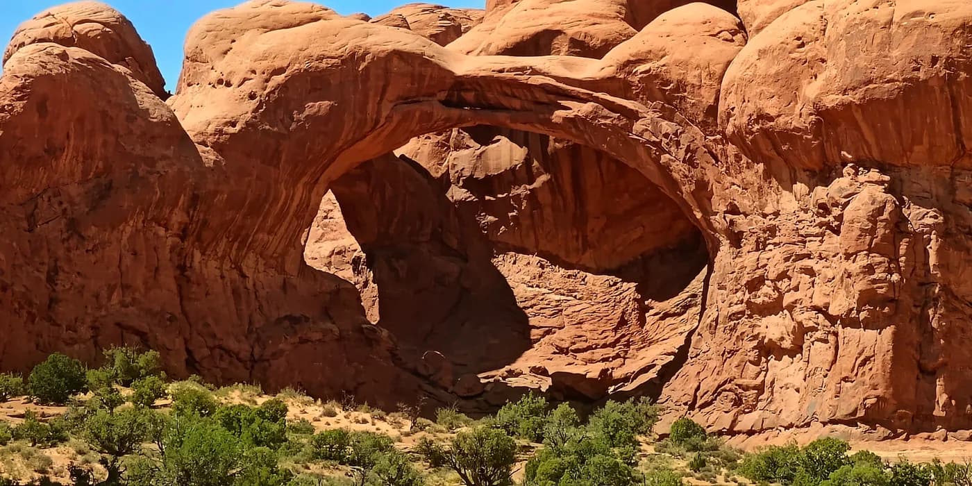Double Arch spans orange sandstone above shrubs and broken rock in Arches National Park.
