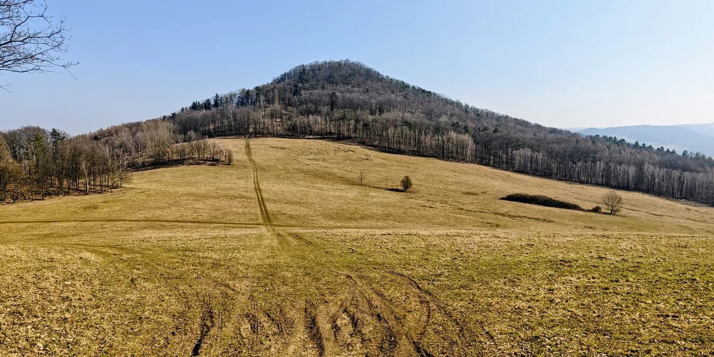 Lotarův vrch above a broad dry meadow, with leafless forest on its slopes under a clear blue sky.