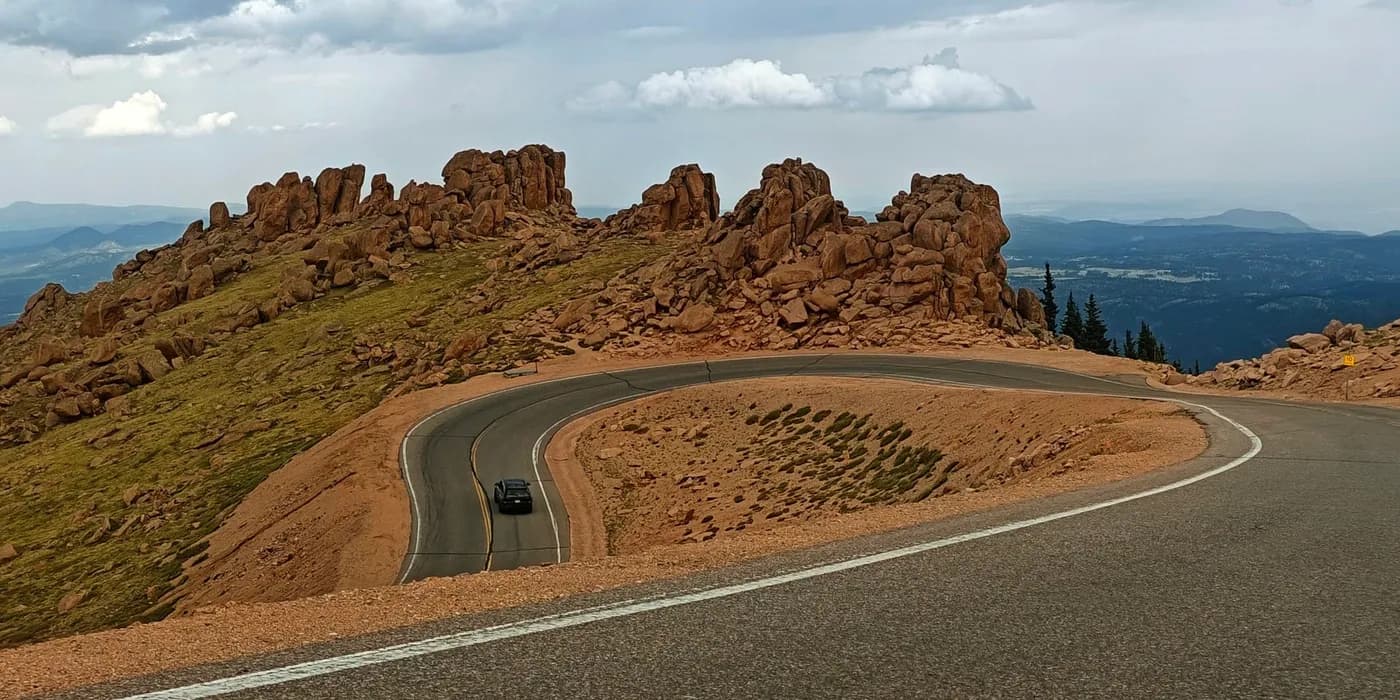 Car rounding a broad hairpin on Pikes Peak Highway through rocky alpine terrain beneath cloudy skies.