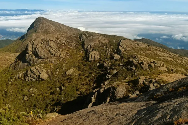 A rounded rocky peak rises above a thick layer of white cloud, with a small yellow trail marker in the foreground.