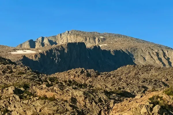 The broad sunlit granite face of Cloud Peak rises above rocky alpine talus under a clear blue sky.
