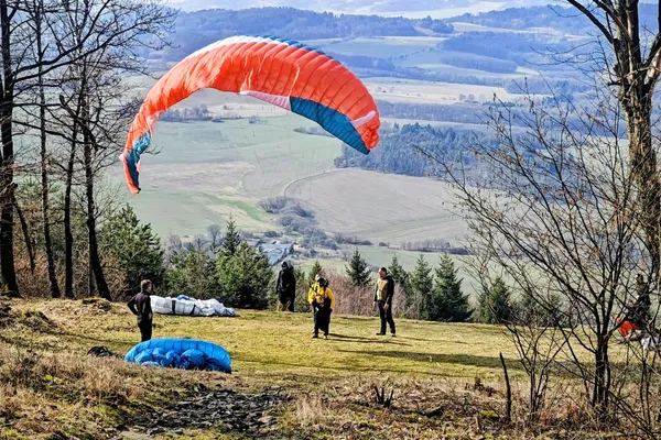 Paragliders on a grassy launch below Doubrava, with an orange canopy overhead and fields in the distance.