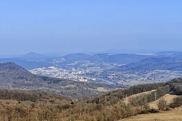Wide view from open summit pasture across wooded hills, scattered settlements and a distant conical peak.
