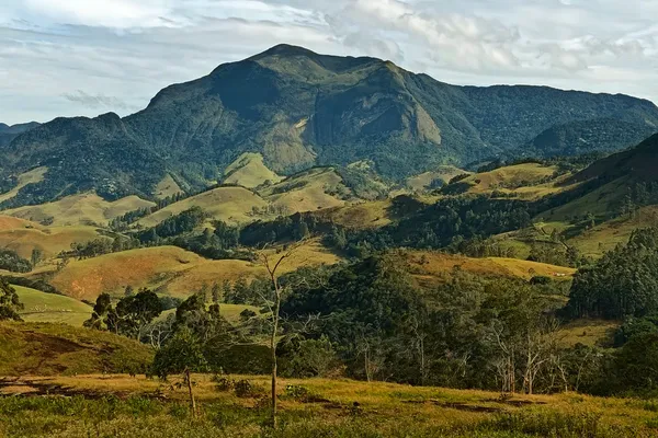 Dark rocky mountain ridge above green valleys and pasture under broken cloud.