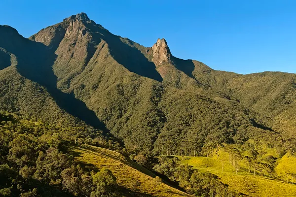 Rugged Pico dos Marins massif above forested slopes under a clear blue sky.