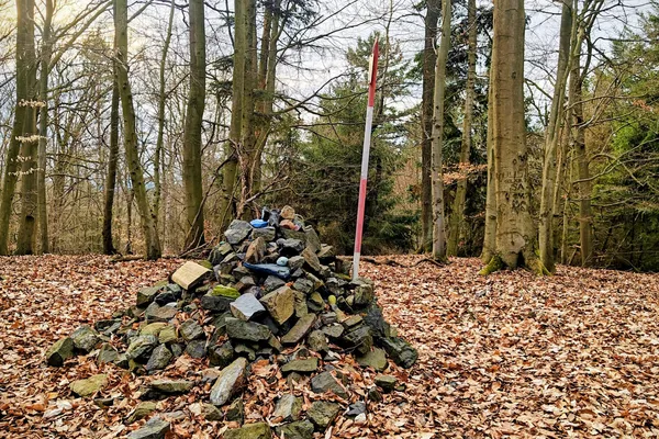 Stone cairn and red-and-white survey pole on the broad wooded summit of Stramchyně in early spring.
