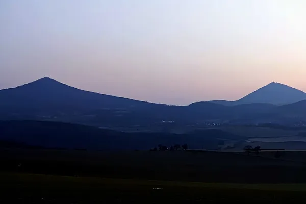 Two dark conical hills silhouetted against a pale pink and blue evening sky.