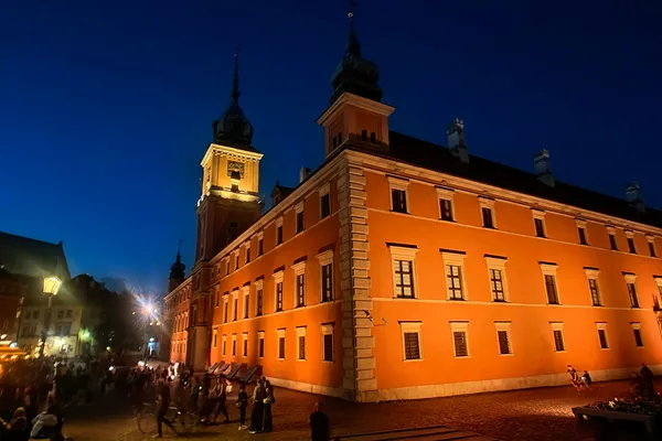 Illuminated Royal Castle on Castle Square in Warsaw at blue hour with people gathered nearby.