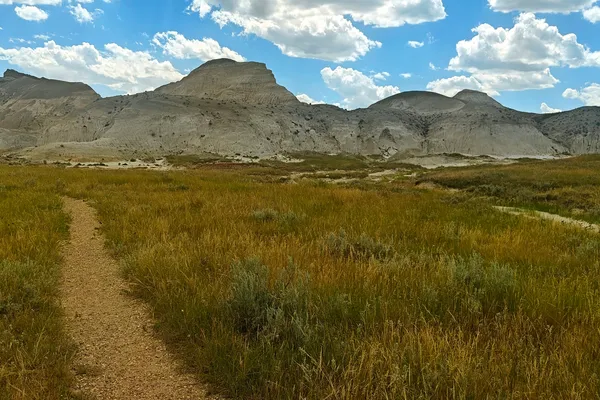 A narrow trail crosses open prairie towards the pale, eroded slopes of White Butte beneath cumulus clouds.